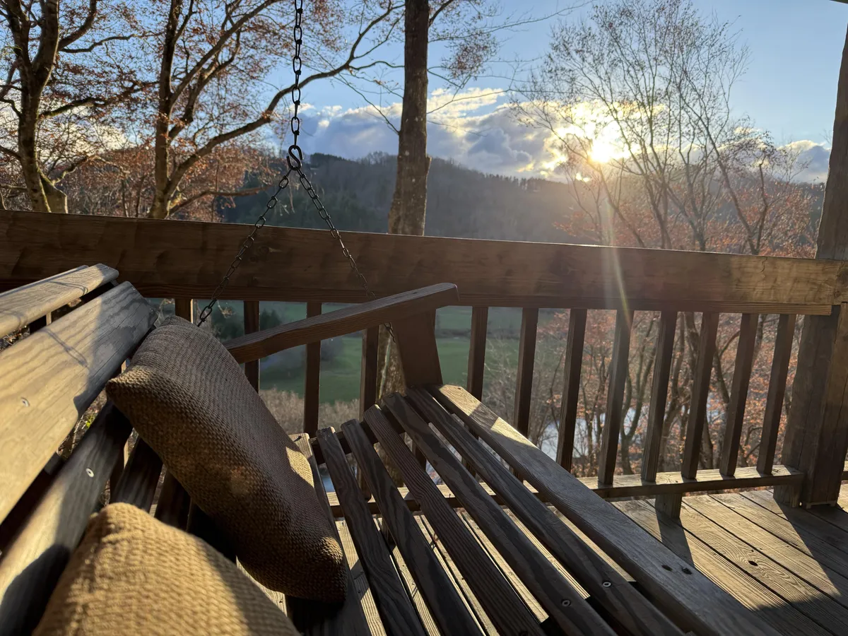 A tranquil exterior scene features a swinging bench overlooking a beautiful sunset, surrounded by trees. The setting oozes relaxation, perfect for quiet moments like morning coffee, meditating, or unwinding with a good book while soaking in natural views.