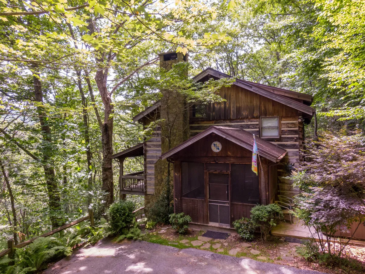 A charming log cabin with a stone chimney nestled among lush greenery, featuring a welcoming screened porch adorned with a flag. The setting suggests relaxation after a day outdoors, enjoying nature walks, gathering on the porch, or sharing stories around a fire pit.