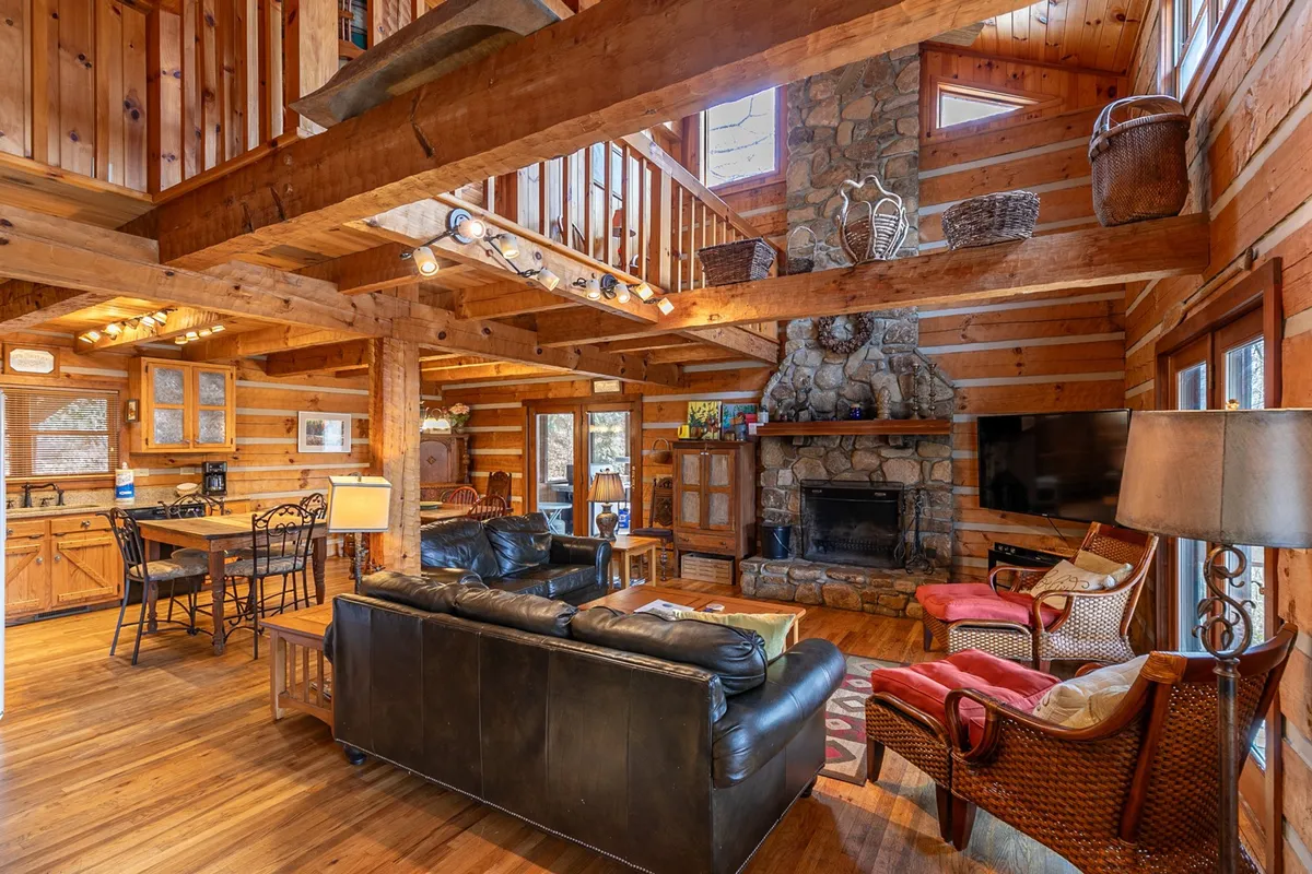 This image showcases a spacious interior with a grand stone fireplace, timbered wooden beams, a large comfortable seating area, and a dining table in the background. The room radiates warmth and provides an inviting atmosphere for gatherings, relaxation, meals, and fireside conversations.
