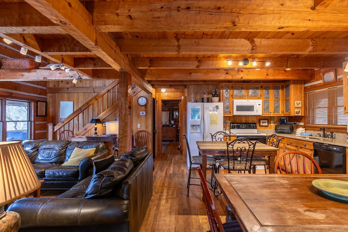A bright, welcoming space with a prominent wooden dining table, plush sofa seating, kitchen appliances, and rustic wooden beams overhead. The arrangement suggests a lively area for both feasts and relaxed hangouts.