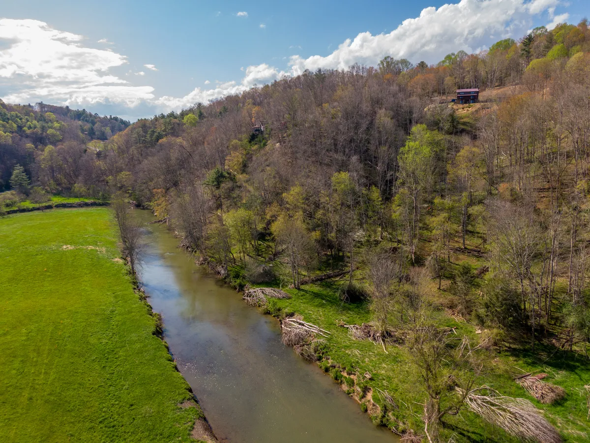 A scenic view of the New River winding through a green meadow, bordered by a tree line and dotted with hillside cabins. The atmosphere is serene, with opportunities for fishing, picnicking, hiking, and birdwatching along the water's edge. Sunset hues add a magical touch to the riverscape, with hints of local wildlife and tranquil outdoor enjoyment.