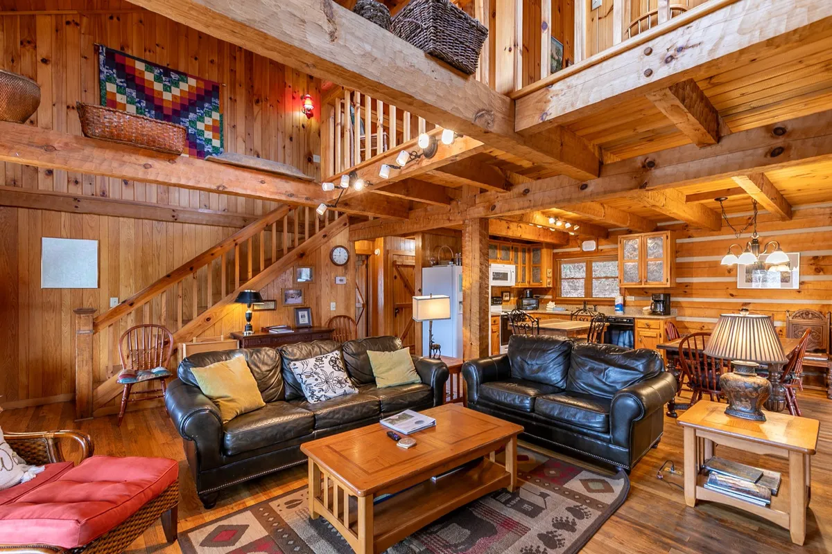 A wide-angle interior view featuring leather sofas, exposed wooden beams, a visible staircase, an open kitchen area, and a large dining table. The open layout seamlessly connects the living, kitchen, and dining spaces, with natural light highlighting the inviting communal zones.