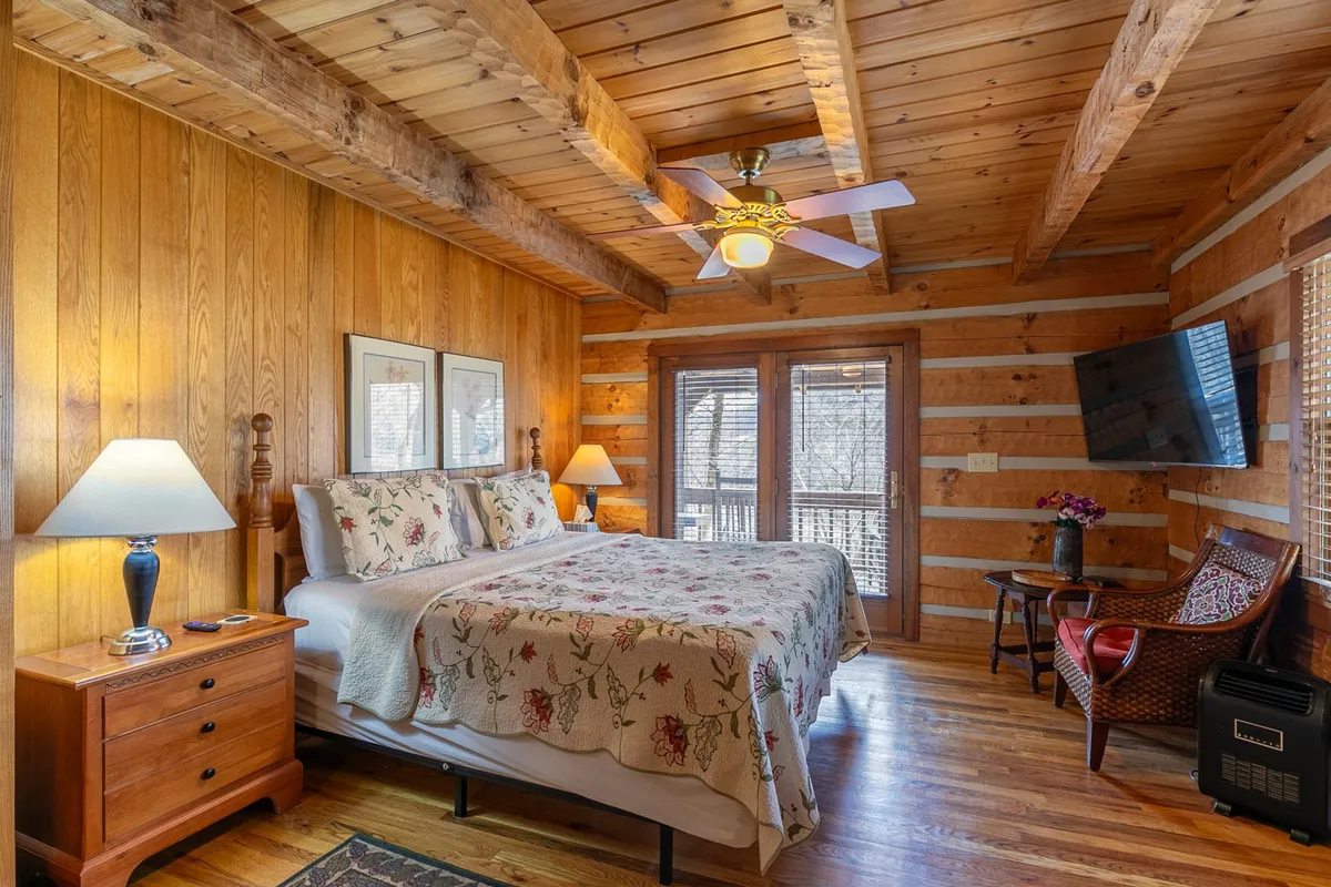 A bright and inviting bedroom featuring a bed with floral bedding, wooden furniture, and a window that lets in natural light. The room exudes warmth and offers a relaxing spot for reading, watching TV, or simply unwinding while enjoying the view outside.