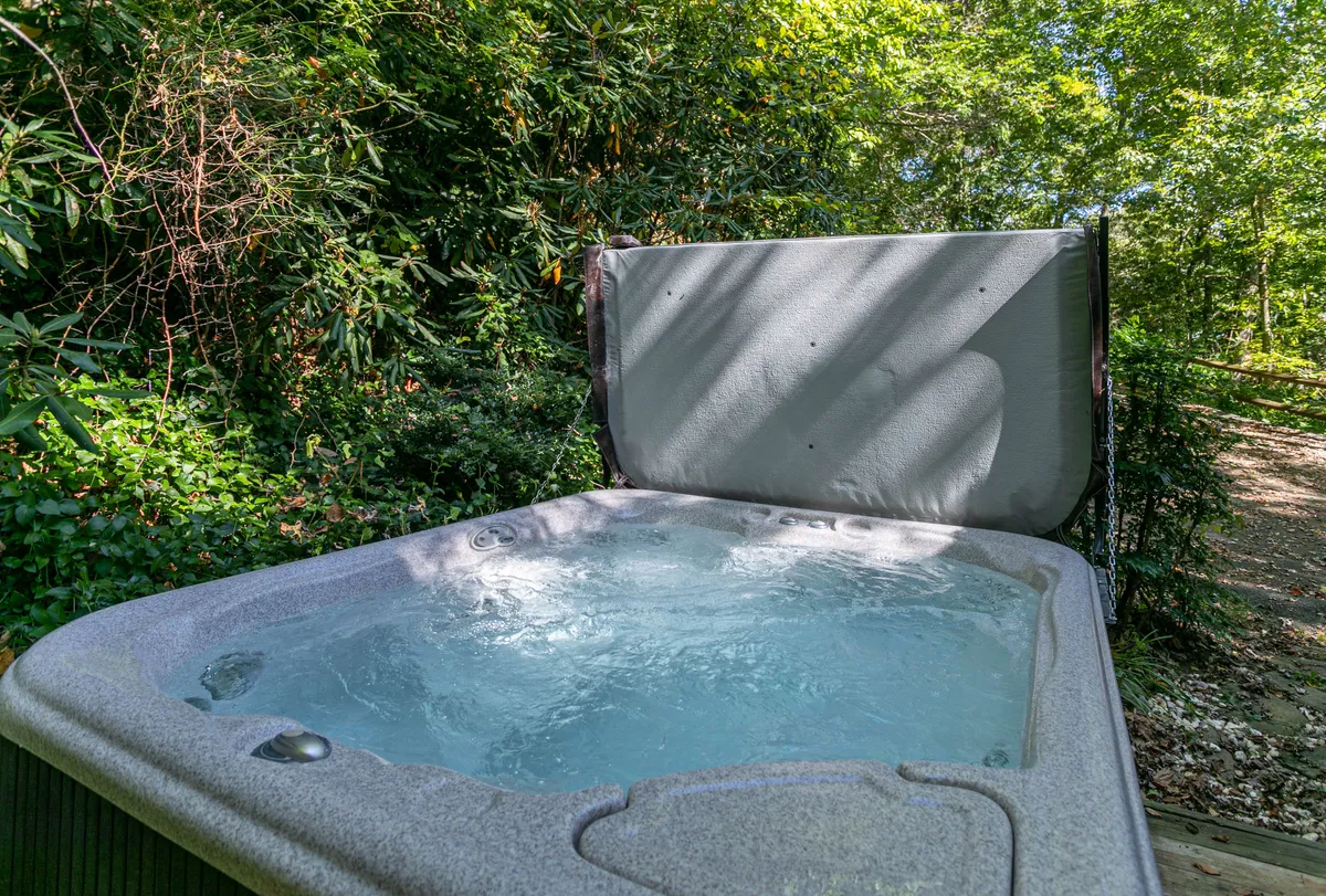 A hot tub nestled on a deck surrounded by lush greenery, with a calm water surface visible. The area is set up for relaxation and socializing, with the natural setting enhancing the sense of privacy and tranquility.