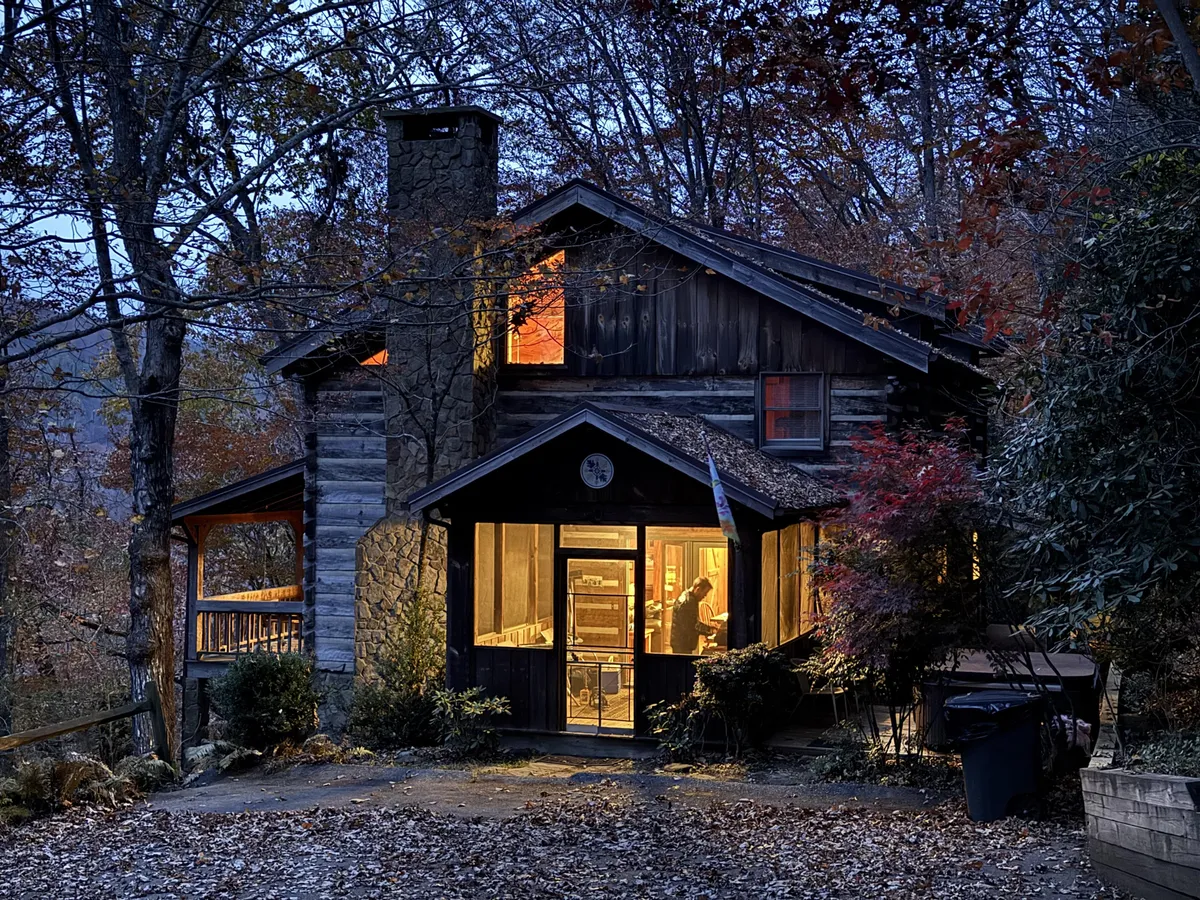 The image shows a classic log cabin perched on a mountain ridge, its warm, glowing windows inviting you in as evening falls. A sturdy stone chimney hints at cozy fires inside, while a welcoming porch offers breathtaking views of the surrounding Blue Ridge Mountains. The setting is tranquil, framed by nature and the promise of outdoor adventures.