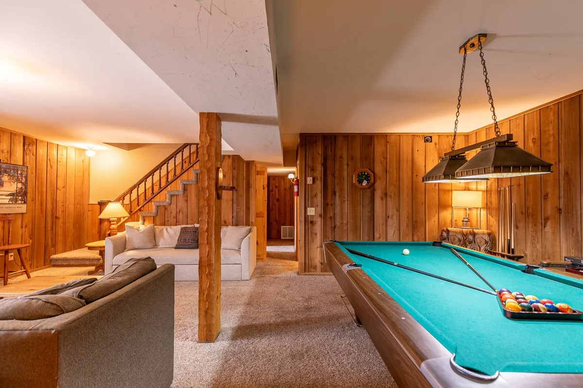 This image features a pool table as the central element, with a sofa nearby, a wooden staircase, and wall décor. The scene gives a sense of casual comfort and friendly competition, inviting guests to play pool or relax together in a spacious recreation room.