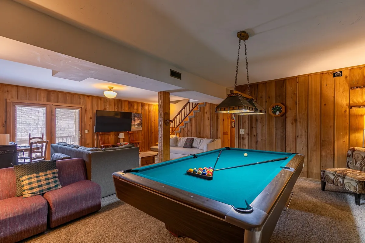 The image displays a pool table, a cozy sofa, a TV, wood paneling, and a staircase, capturing the full range of entertainment options available in the recreation room. The inviting atmosphere is ideal for both movie nights and pool tournaments.